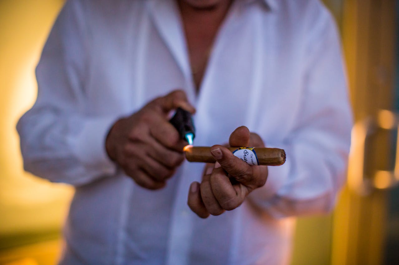Man in white shirt lighting a cigar with a torch lighter, cigar band visible, warm ambient lighting